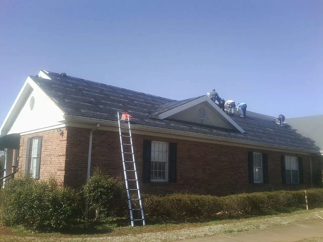 Crew tearing shingles off a brick ranch roof with a ladder set — Emergency Roof Repair work by our crew in Pilot Mountain