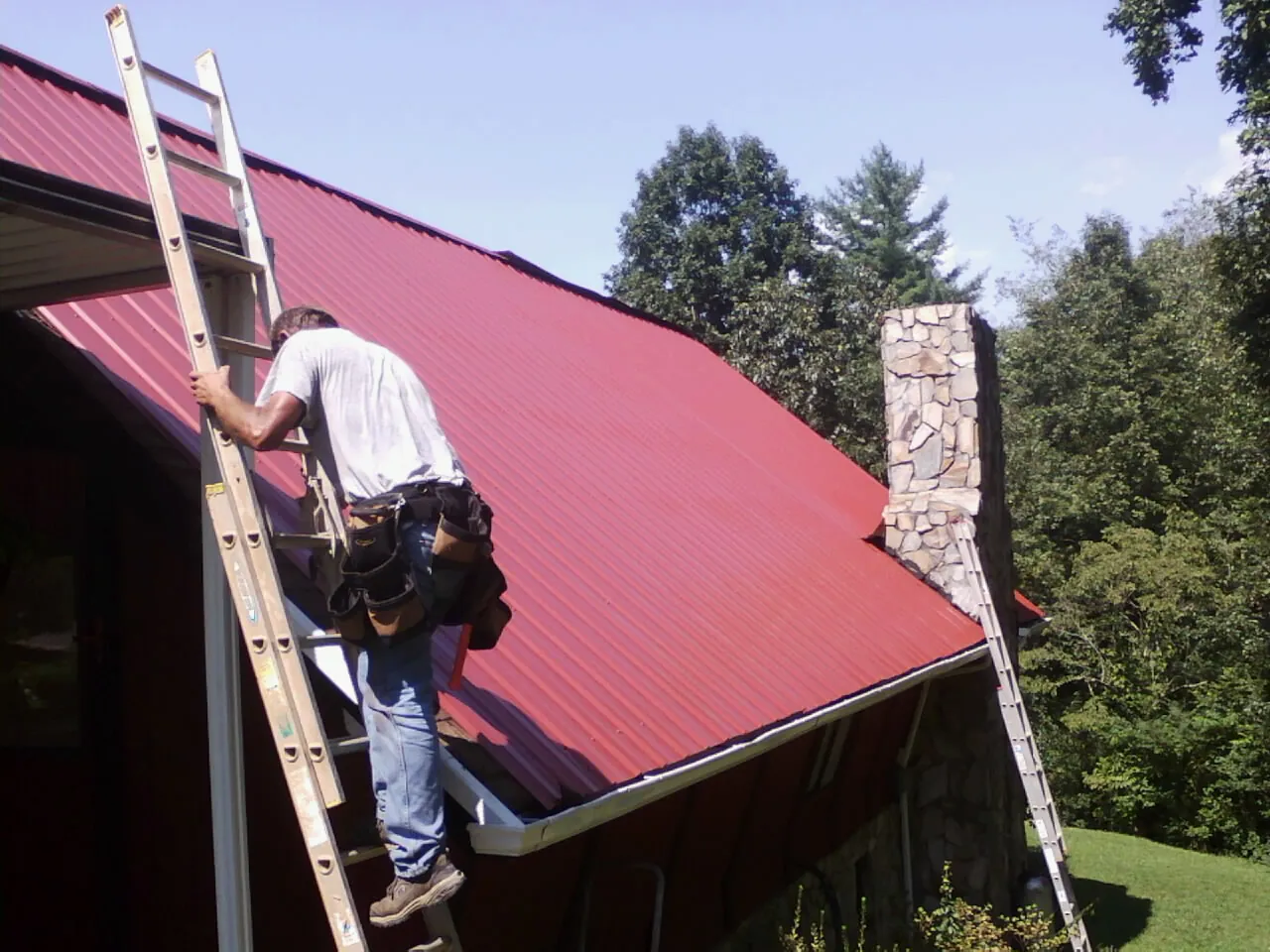 Roofer on a ladder beside a red metal roof and stone chimney — Soffit & Fascia Repair by Roofing Brothers in Pilot Mountain